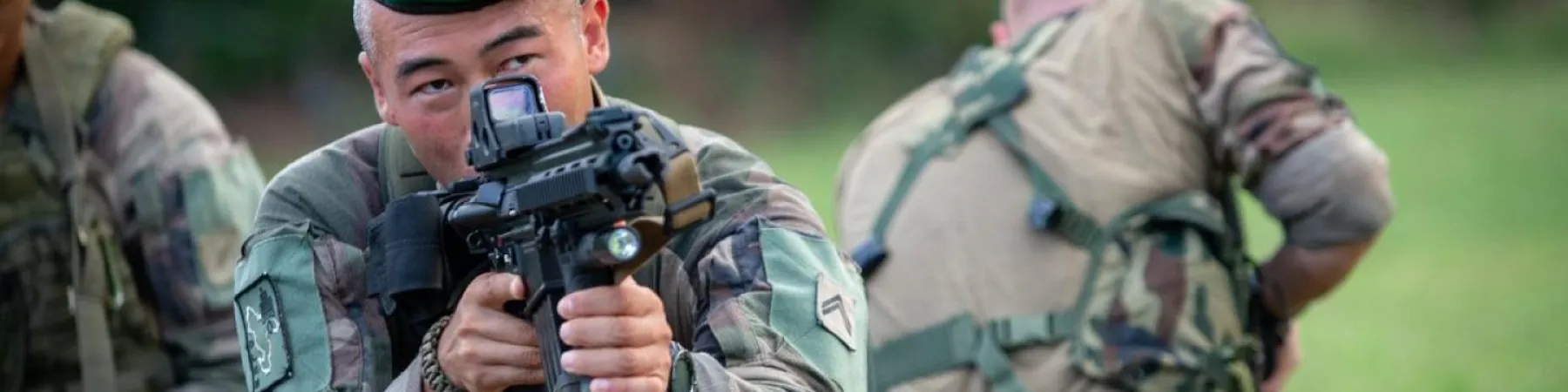 Soldat visant avec un fusil d'assaut équipé d'une lunette, en position de tir. Un autre soldat est visible dans le flou à l'arrière-plan.